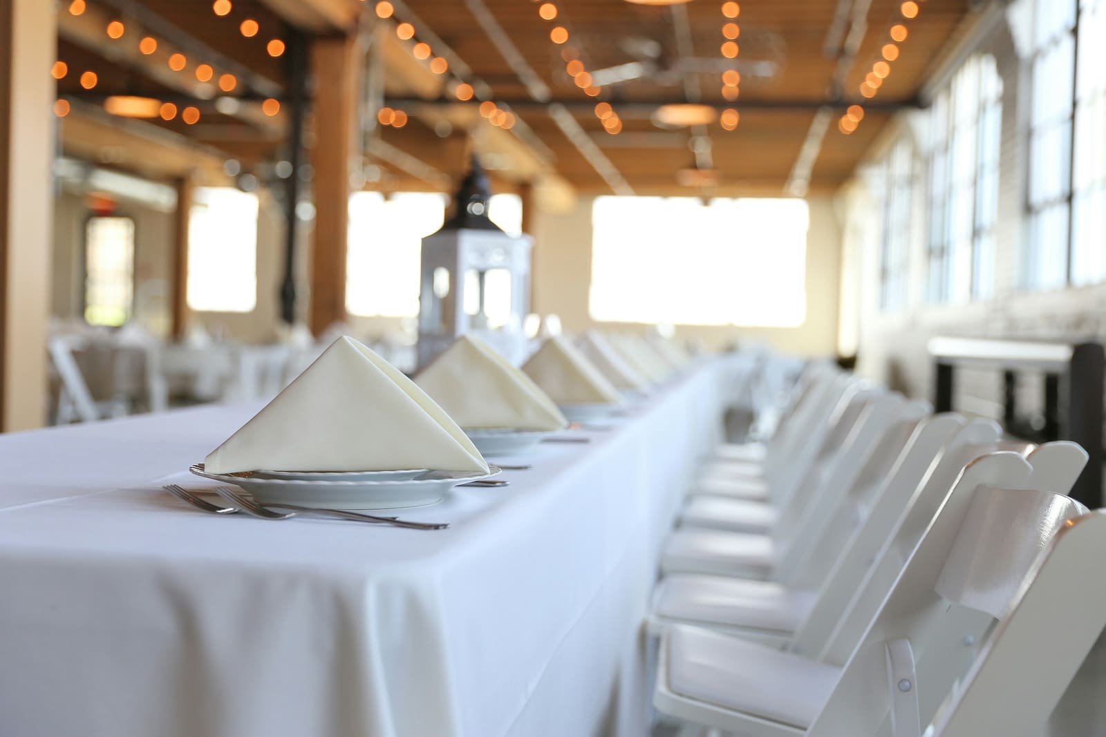 Long reception table close-up: folded white napkins on plates with bistro lights overhead