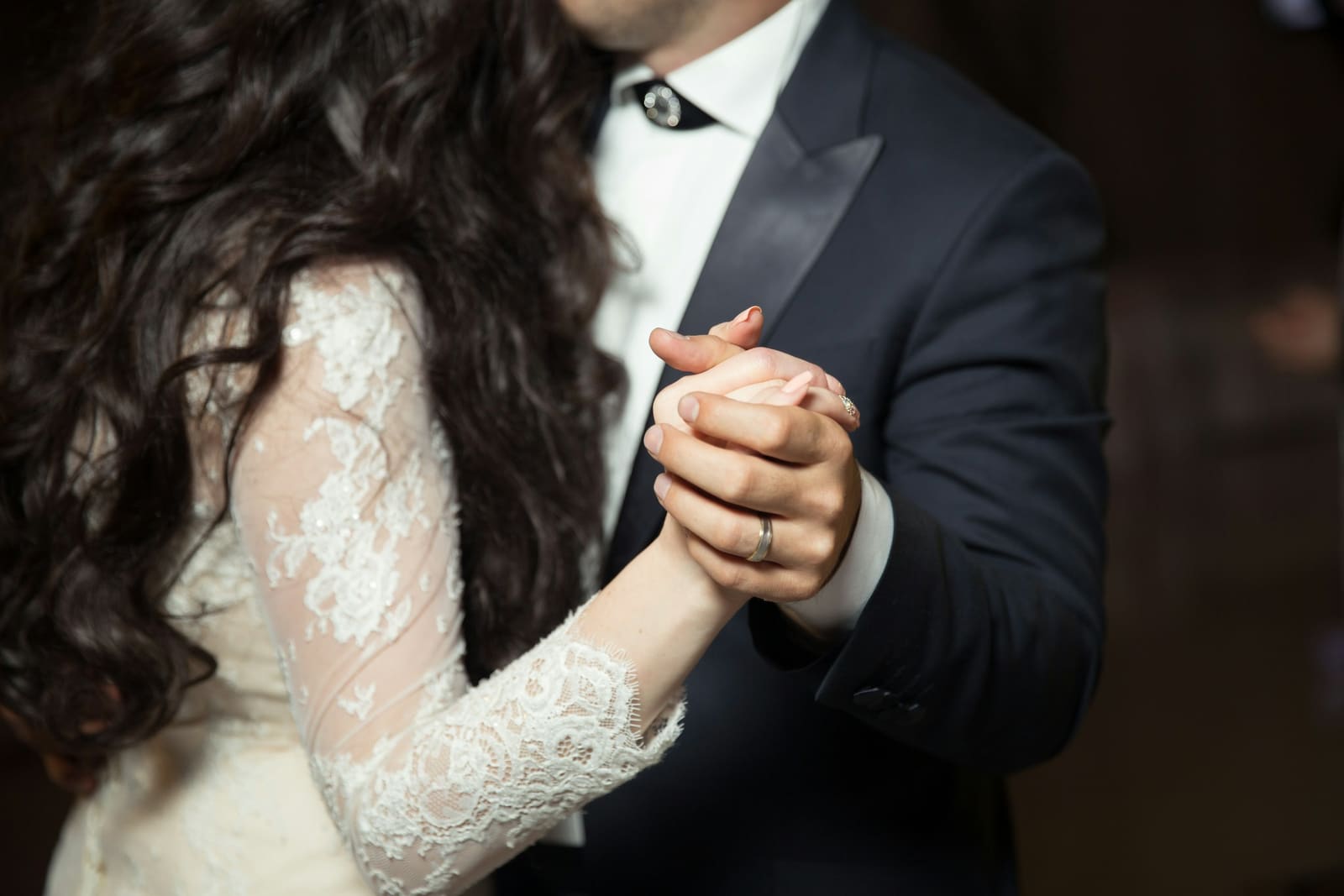 First dance close-up: bride in lace gown and groom in tuxedo with clasped hands